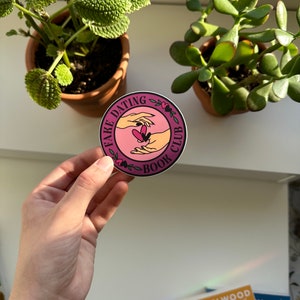 May include: A round sticker with a pink background and white text that reads "Fake Dating Book Club". The sticker features a graphic of two hands reaching towards each other with a heart in the middle.