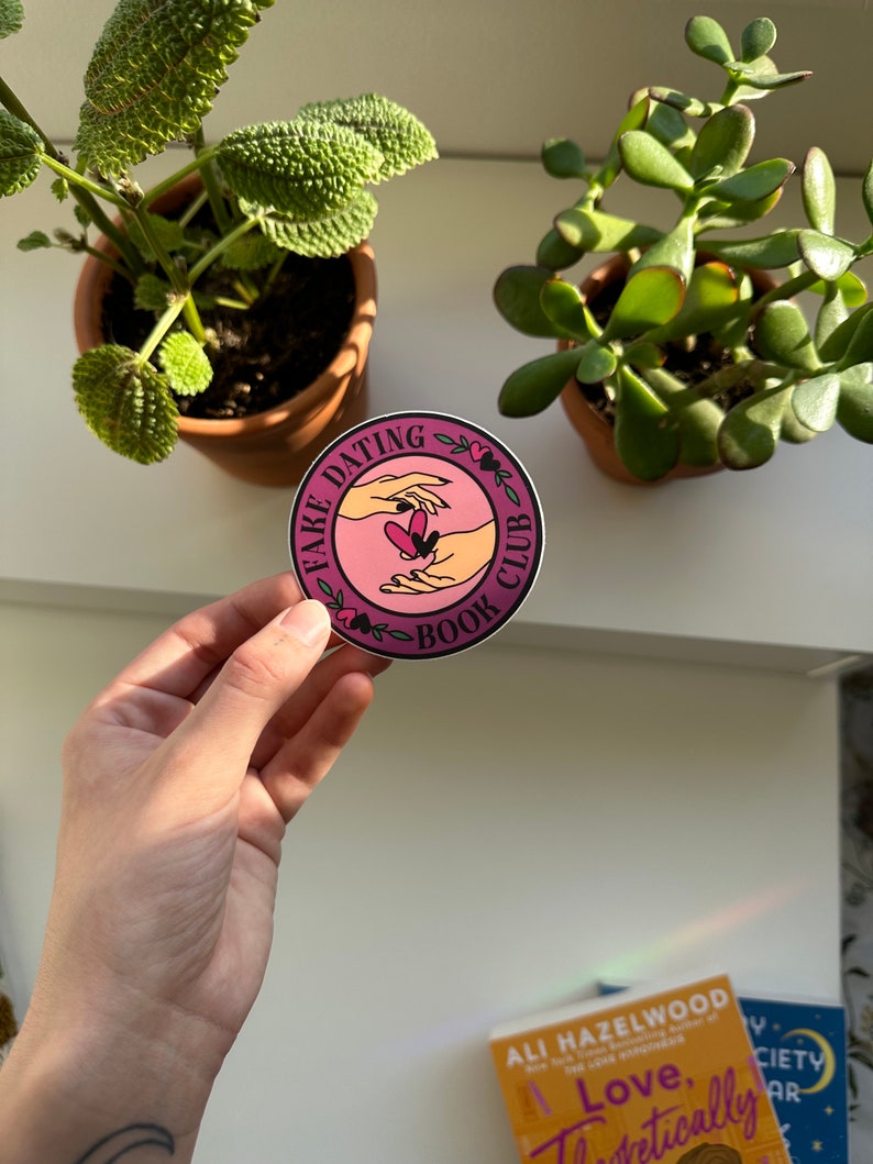 May include: A round sticker with a pink background and white text that reads "Fake Dating Book Club". The sticker features a graphic of two hands reaching towards each other with a heart in the middle.