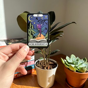 May include: A tarot card with a purple and blue fairy sitting on a book. The card is titled "The Romantasy" and has a crescent moon and stars in the background.