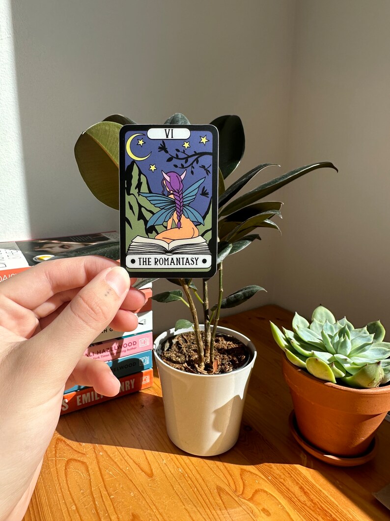 May include: A tarot card with a purple and blue fairy sitting on a book. The card is titled "The Romantasy" and has a crescent moon and stars in the background.