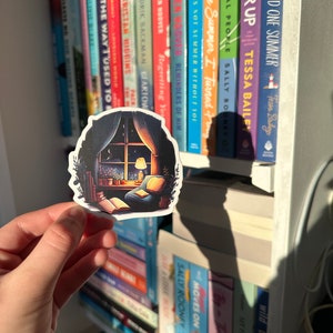 May include: A sticker featuring a cozy reading nook scene with a window, armchair, and open book. The sticker has a dark border and is held in front of a bookshelf filled with colorful book spines. The sticker's design includes a warm lamp and a view of the night sky.