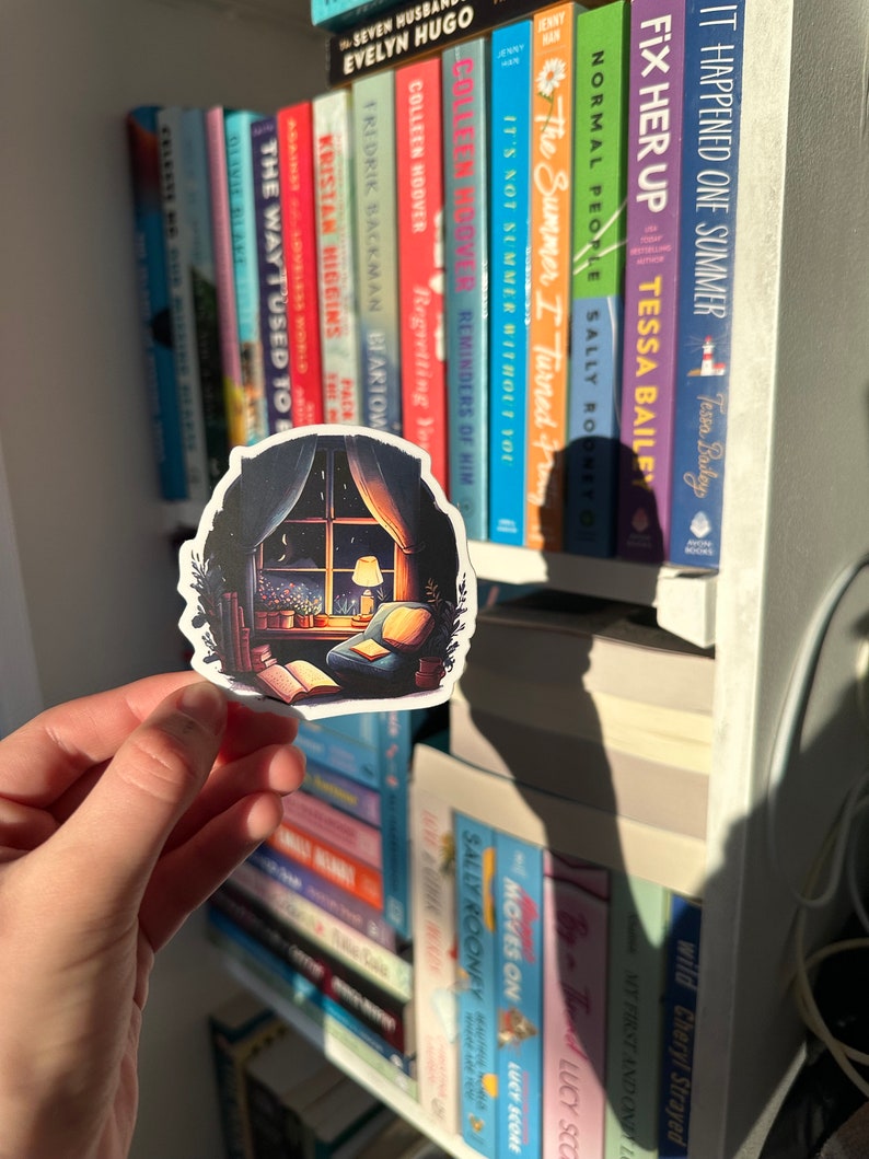 May include: A sticker featuring a cozy reading nook scene with a window, armchair, and open book. The sticker has a dark border and is held in front of a bookshelf filled with colorful book spines. The sticker's design includes a warm lamp and a view of the night sky.