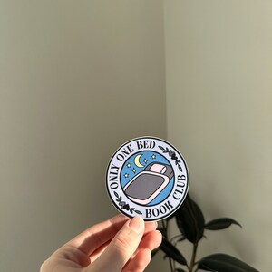 May include: A round sticker with a blue background and white text that reads "Only One Bed Book Club". The sticker features a black and white illustration of a bed with a pillow and a crescent moon with stars above it.