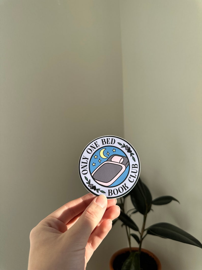 May include: A round sticker with a blue background and white text that reads "Only One Bed Book Club". The sticker features a black and white illustration of a bed with a pillow and a crescent moon with stars above it.