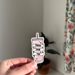 K&ouml;nnte beinhalten: Rosafarbener Aufkleber mit einem rosa Getr&auml;nk und dem Text "Finish Your TBR" in Schwarz.