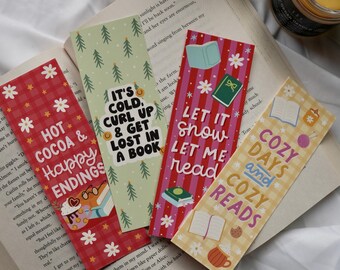 May include: Four colorful bookmarks with winter-themed designs and text, including "Hot Cocoa & Happy Endings" and "It's Cold, Curl Up & Get Lost in a Book." The bookmarks are on an open book.