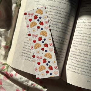Pastel bookmark with a repeating pattern of croissants, strawberries, blueberries, flowers, bows, and hearts on an open book with soft floral bedding and natural sunlight