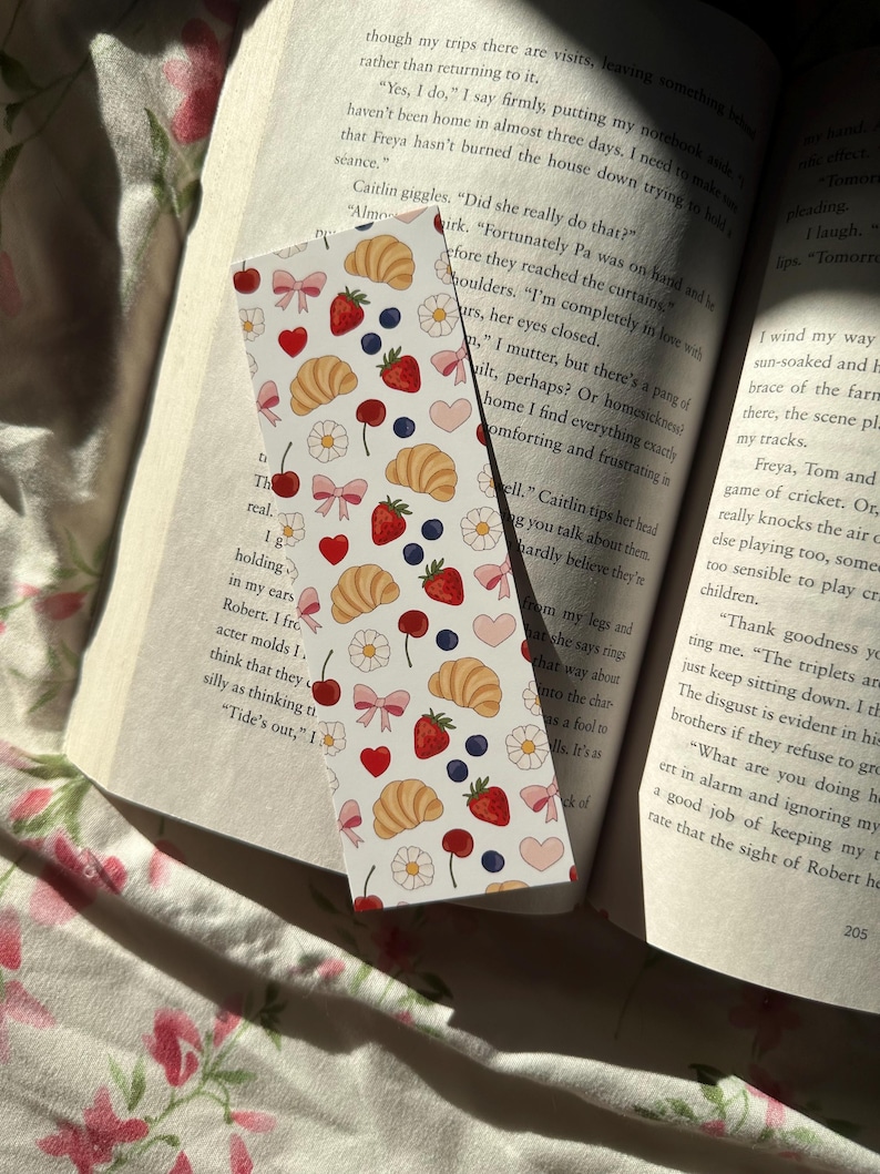 Pastel bookmark with a repeating pattern of croissants, strawberries, blueberries, flowers, bows, and hearts on an open book with soft floral bedding and natural sunlight