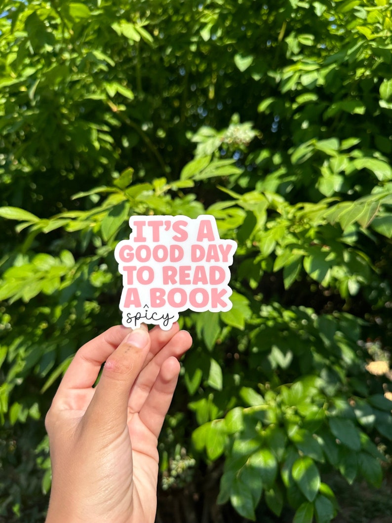 May include: A pink and white sticker with the text "It's a good day to read a book" and the word "spicy" underneath. The sticker is being held in front of a green leafy background.