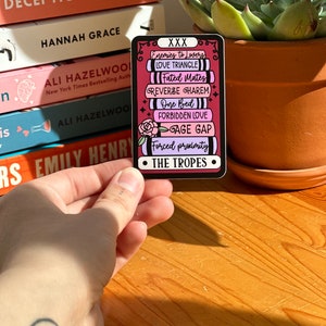 May include: A black and white sticker with the text "XXX The Tropes" and a list of romantic tropes including "Enemies to Lovers", "Love Triangle", "Fated Mates", "Reverse Harem", "One Bed", "Forbidden Love", "Age Gap", and "Forced Proximity".