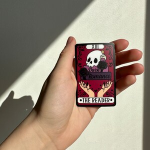 May include: A tarot card with a black and white skull, a candle, and two hands with long nails. The card is red with black trim and the text "Dark Romance" and "The Reader" in black. The card is numbered "XIII".