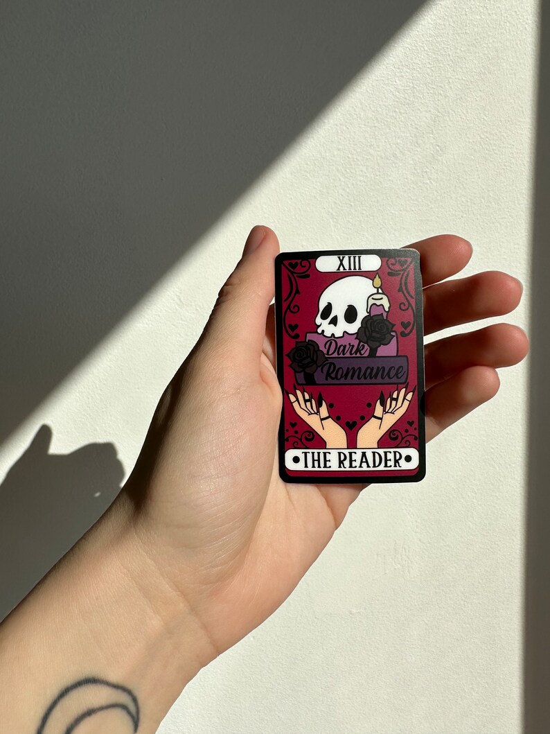 May include: A tarot card with a black and white skull, a candle, and two hands with long nails. The card is red with black trim and the text "Dark Romance" and "The Reader" in black. The card is numbered "XIII".