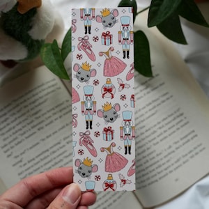 Puede incluir: Marcapáginas rectangular con un patrón repetitivo de ilustraciones temáticas de ballet sobre un fondo blanco. El diseño incluye cascanueces, ratones, zapatillas de ballet, vestidos y cajas de regalo en rosa, azul y rojo.