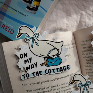 May include: A white goose bookmark with a blue bow and purse, with the text "ON MY WAY TO THE COTTAGE". The bookmark is placed on an open book with text.