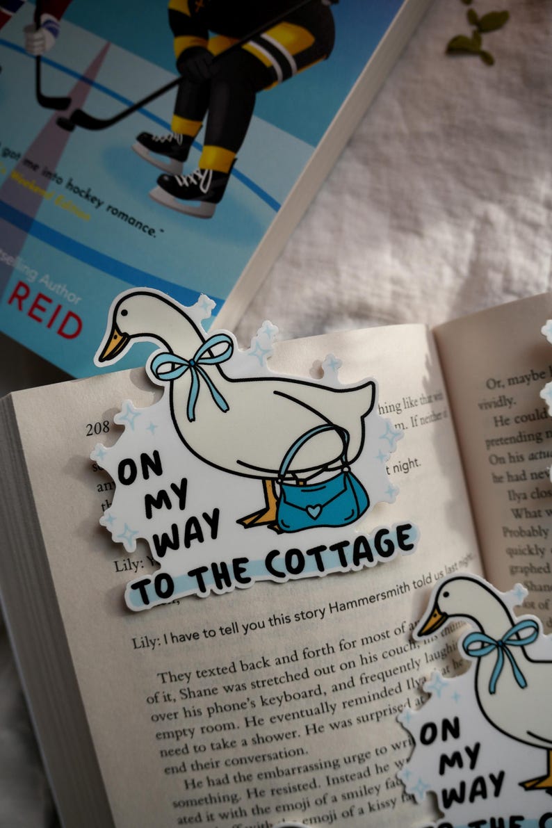 May include: A white goose bookmark with a blue bow and purse, with the text "ON MY WAY TO THE COTTAGE". The bookmark is placed on an open book with text.