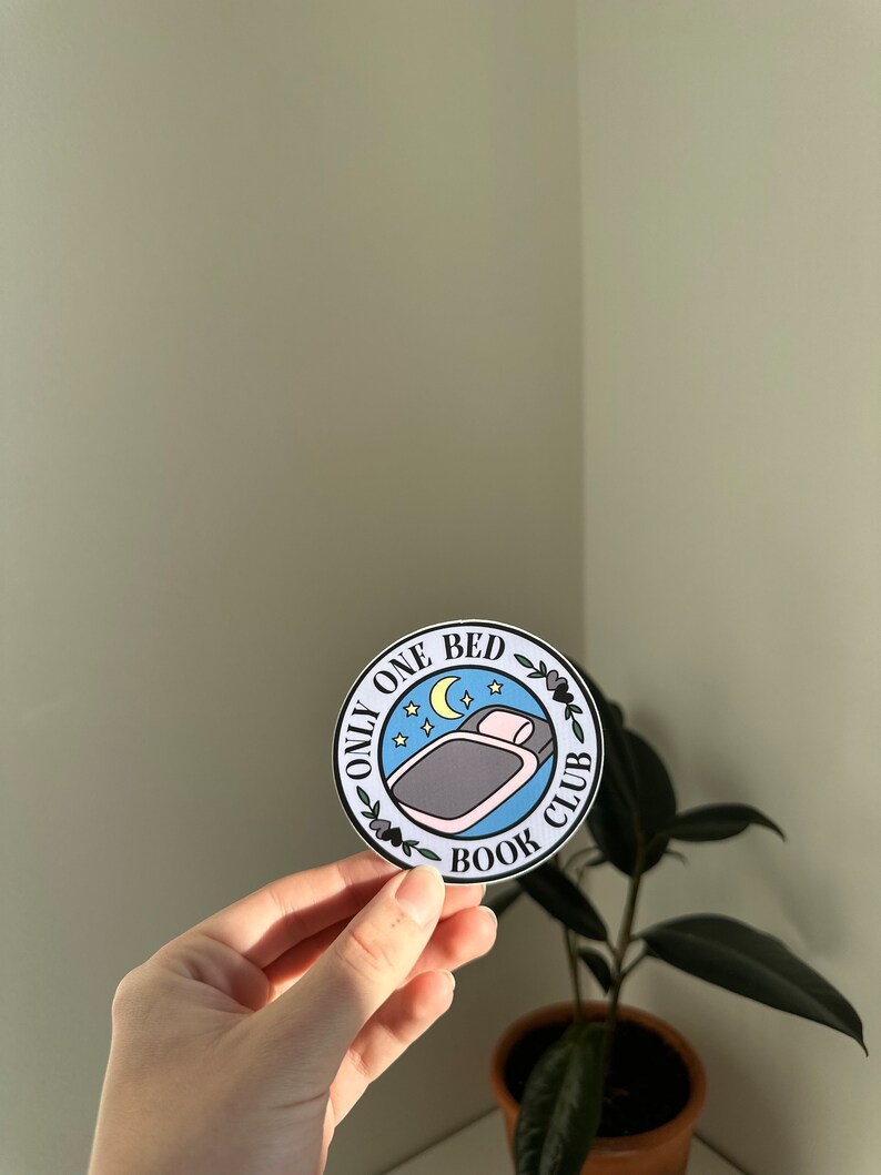 May include: A blue and white round sticker with the text "Only One Bed Book Club" and a cartoon illustration of a bed with a pillow and a crescent moon.