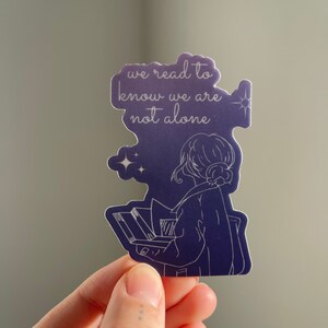 May include: A purple and white sticker with a line drawing of a person reading a book. The text on the sticker reads "we read to know we are not alone".