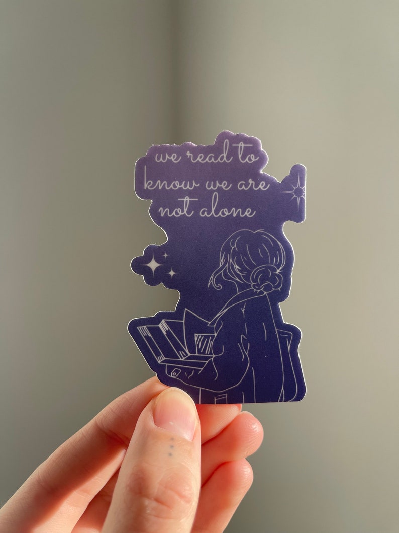 May include: A purple and white sticker with a line drawing of a person reading a book. The text on the sticker reads "we read to know we are not alone".