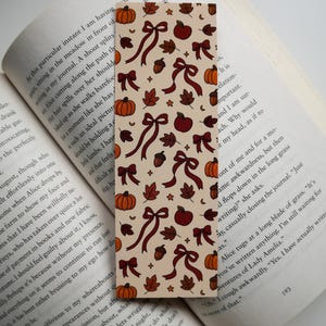 Puede incluir: Un marcapáginas rectangular con un fondo beige, con un patrón repetido de lazos marrones, calabazas, manzanas, bellotas y hojas. El marcapáginas está colocado en un libro abierto.
