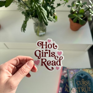 May include: A white sticker with the text "Hot Girls Read" in maroon lettering and pink hearts. The sticker is being held by a hand.