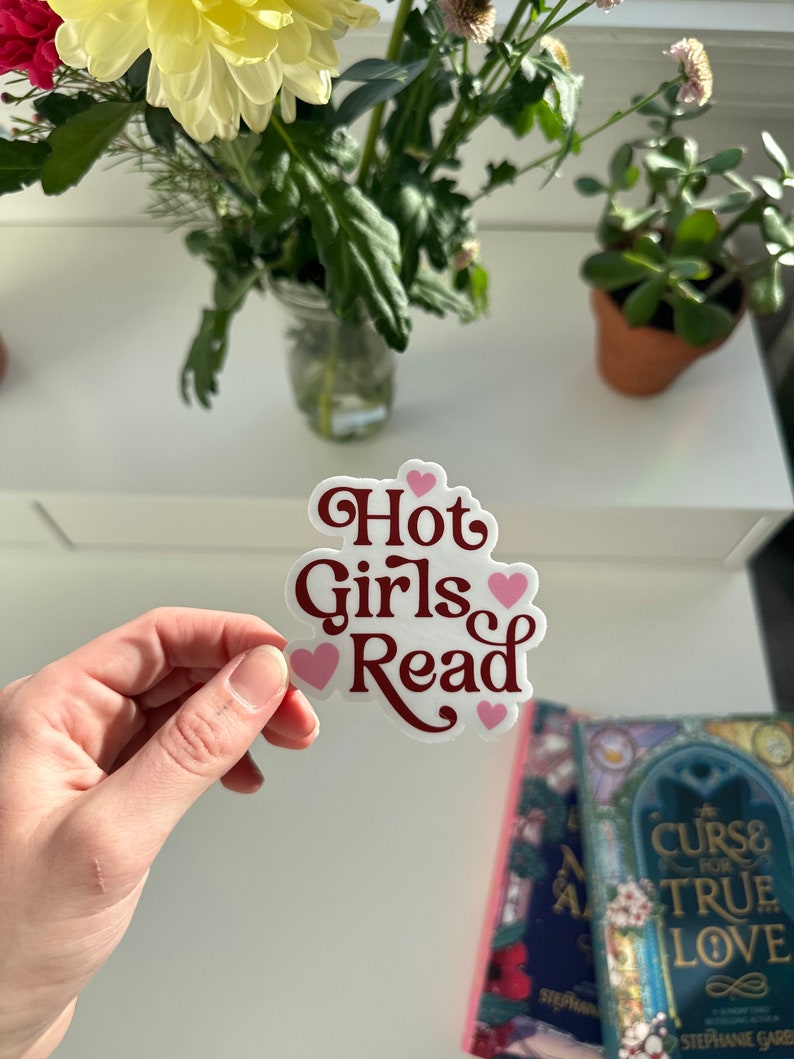May include: A white sticker with the text "Hot Girls Read" in maroon lettering and pink hearts. The sticker is being held by a hand.
