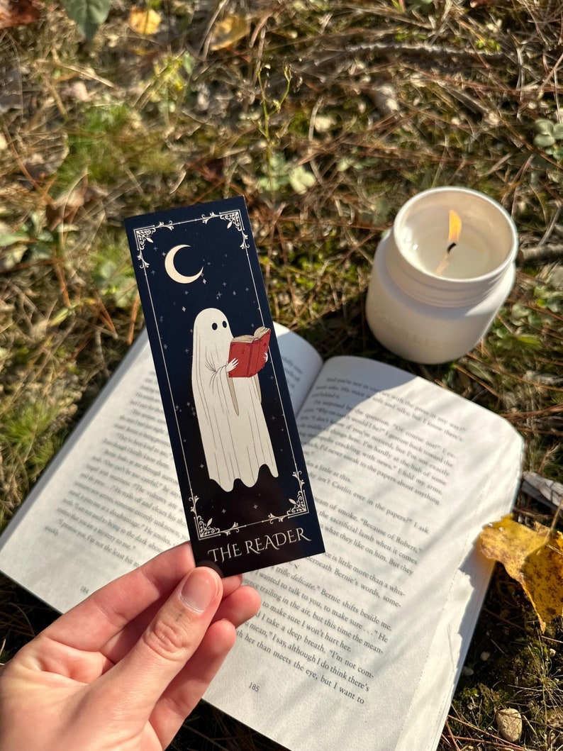May include: A black bookmark with a white ghost reading a red book. The bookmark has a crescent moon and stars in the background. The text "THE READER" is printed on the bookmark.