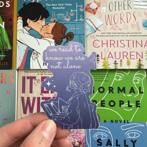 May include: A purple sticker with a silhouette of a person reading a book. The sticker has the text "we read to know we are not alone" on it. The sticker is placed on a bookshelf with several books visible.
