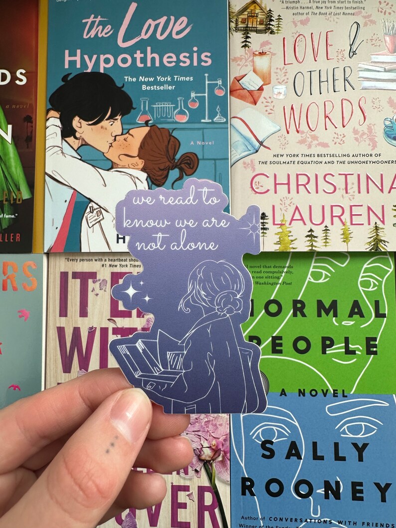 May include: A purple sticker with a silhouette of a person reading a book. The sticker has the text "we read to know we are not alone" on it. The sticker is placed on a bookshelf with several books visible.