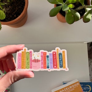 May include: A sticker of a bookshelf with a variety of books, each with a different title related to love and relationships. The books are in a rainbow of colors and have a whimsical, cartoonish style. The sticker features a small pot of flowers with white blossoms.