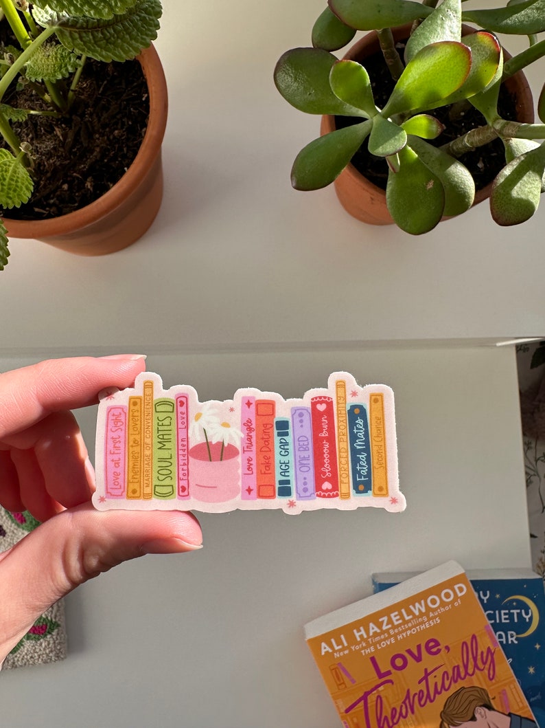 May include: A sticker of a bookshelf with a variety of books, each with a different title related to love and relationships. The books are in a rainbow of colors and have a whimsical, cartoonish style. The sticker features a small pot of flowers with white blossoms.
