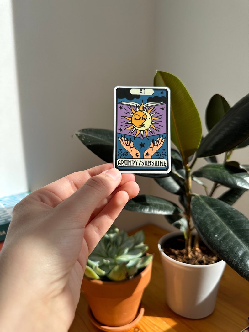 May include: A tarot card with a purple background and a sun and moon design. The card is labeled "XI" and "GRUMPY/SUNSHINE".