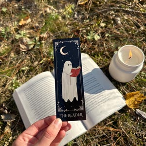 May include: A black bookmark with a white ghost reading a red book. The bookmark has a crescent moon and stars on a dark blue background. The text "THE READER" is printed at the bottom of the bookmark.