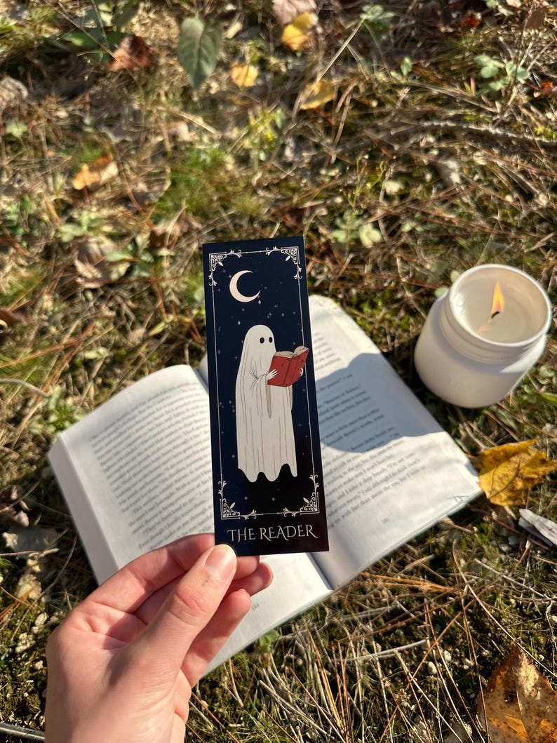 May include: A black bookmark with a white ghost reading a red book. The bookmark has a crescent moon and stars on a dark blue background. The text "THE READER" is printed at the bottom of the bookmark.