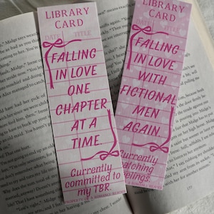 May include: Two pink bookmarks with the text "Falling in Love One Chapter at a Time" and "Falling in Love with Fictional Men Again." The bookmarks have a library card design with a bow and heart details.