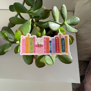 May include: A sticker of a stack of colorful books with titles like "Love at First Sight", "Soul Mates", "Forbidden Love", "Fake Dating", "One Bed", "Slow Burn", and "Fated Mates".