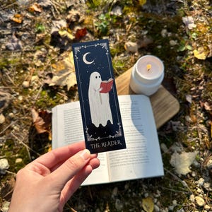 May include: A black bookmark with a white ghost reading a book. The bookmark has a crescent moon and stars in the background. The text "THE READER" is at the bottom of the bookmark.