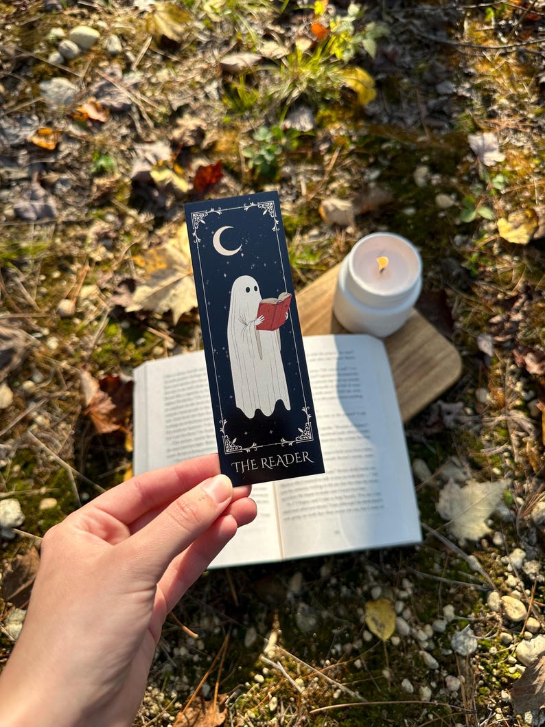 May include: A black bookmark with a white ghost reading a book. The bookmark has a crescent moon and stars in the background. The text "THE READER" is at the bottom of the bookmark.