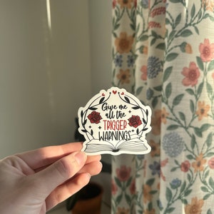 May include: A white sticker with a black and red design. The sticker features a book with the text "Give me all the trigger warnings" in red lettering. The book is surrounded by a wreath of roses and leaves.
