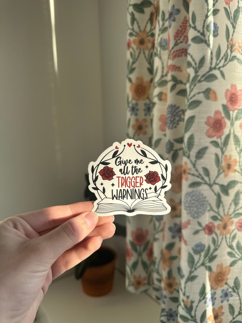 May include: A white sticker with a black and red design. The sticker features a book with the text "Give me all the trigger warnings" in red lettering. The book is surrounded by a wreath of roses and leaves.