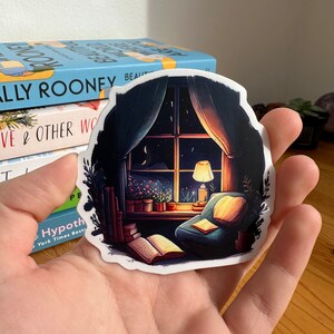 May include: A sticker featuring a cozy reading nook scene. The illustration depicts a window with a night sky view, a lit lamp, a comfortable armchair, and open books. The sticker has a white border and is held in a hand.