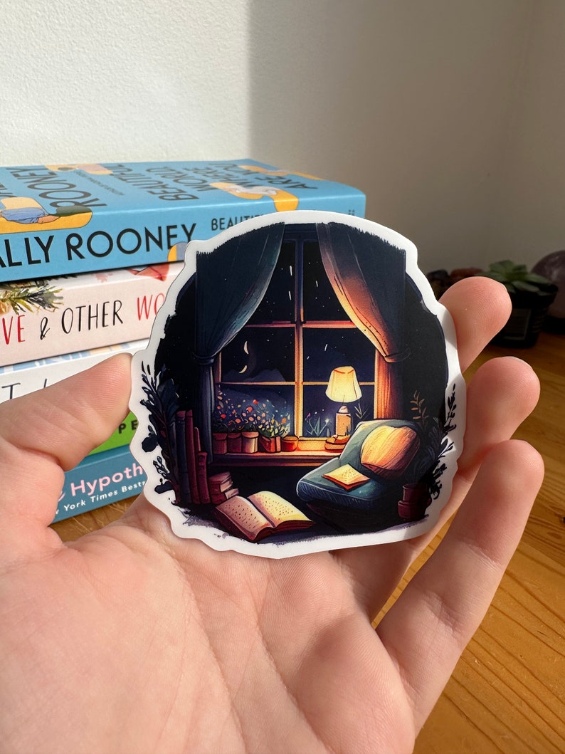 May include: A sticker featuring a cozy reading nook scene. The illustration depicts a window with a night sky view, a lit lamp, a comfortable armchair, and open books. The sticker has a white border and is held in a hand.