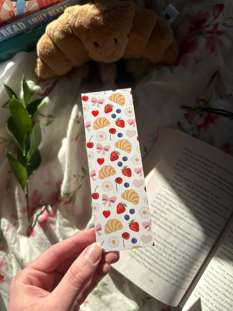 Pastel bookmark with a repeating pattern of croissants, strawberries, blueberries, flowers, bows, and hearts held in front of an open book with soft floral bedding, a croissant stuffed prop and natural sunlight