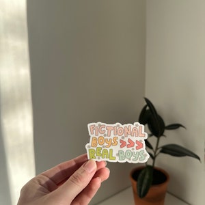 K&ouml;nnte beinhalten: Ein Aufkleber mit dem Text "Fictional Boys >>> Real Boys" in einem Regenbogen-Farbschema mit kleinen Blumenakzenten.