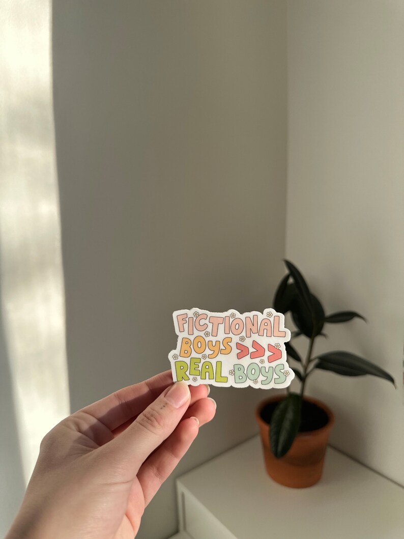 K&ouml;nnte beinhalten: Ein Aufkleber mit dem Text "Fictional Boys >>> Real Boys" in einem Regenbogen-Farbschema mit kleinen Blumenakzenten.