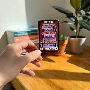 May include: A sticker with a pink and black background featuring a list of common romance tropes, including "Enemies to Lovers", "Love Triangle", "Fated Mates", "Reverse Harem", "One Bed", "Forbidden Love", "Age Gap", and "Forced Proximity". The sticker also includes the text "XXX" and "The Tropes".