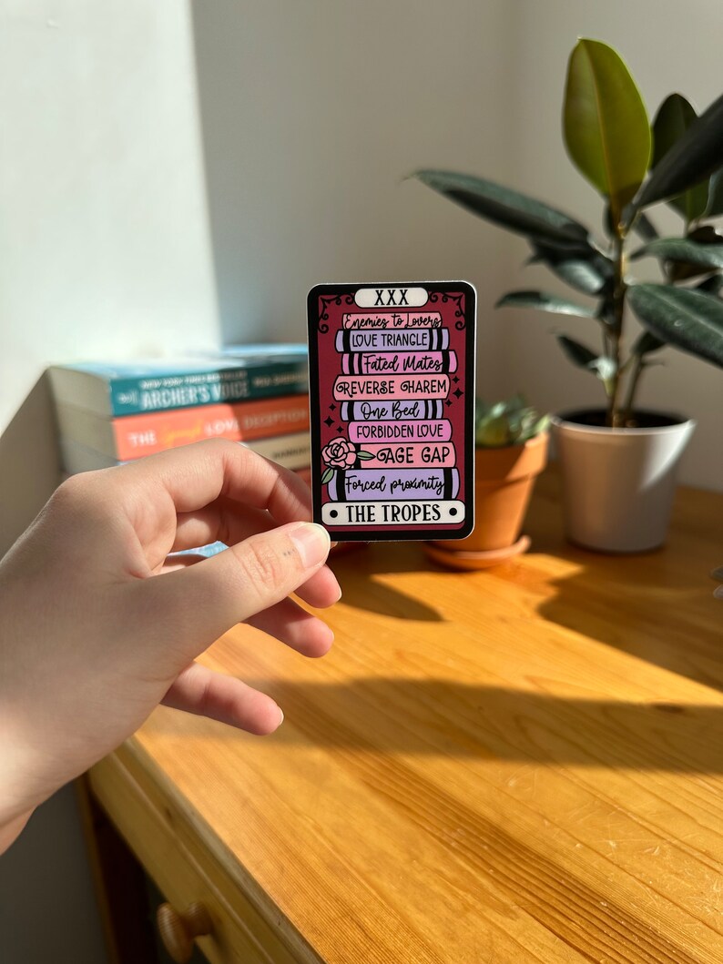 May include: A sticker with a pink and black background featuring a list of common romance tropes, including "Enemies to Lovers", "Love Triangle", "Fated Mates", "Reverse Harem", "One Bed", "Forbidden Love", "Age Gap", and "Forced Proximity". The sticker also includes the text "XXX" and "The Tropes".