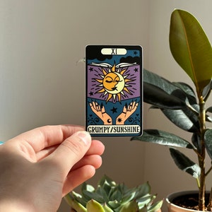 May include: A sticker with a purple background and a sun and moon design. The sun and moon are kissing and there are stars and hearts around them. The text "GRUMPY/SUNSHINE" is at the bottom of the sticker.