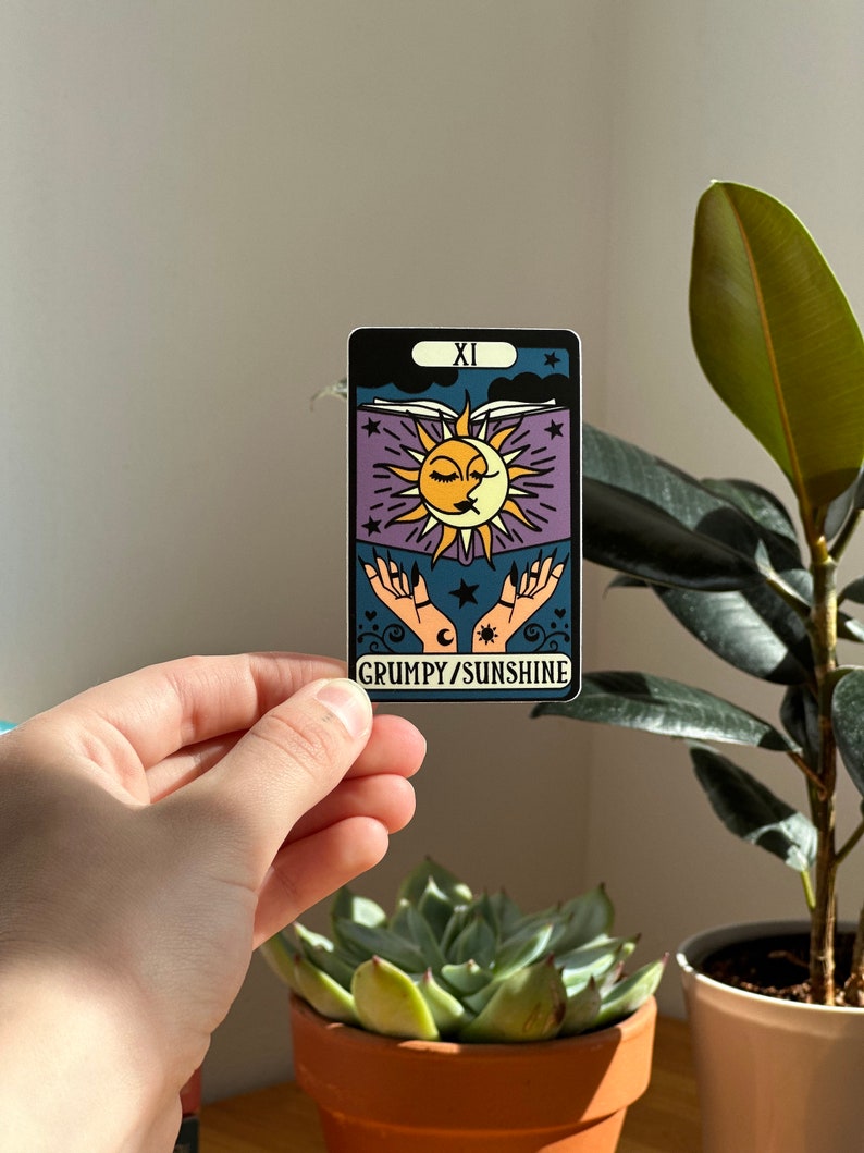 May include: A sticker with a purple background and a sun and moon design. The sun and moon are kissing and there are stars and hearts around them. The text "GRUMPY/SUNSHINE" is at the bottom of the sticker.