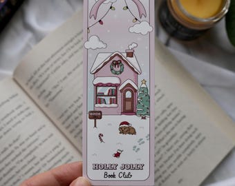 May include: A pink bookmark with a Christmas theme. It features a house with snow, a Christmas tree, and a dog wearing a Santa hat. The text "HOLLY JOLLY Book Club" is at the bottom. A pink bow and string lights are at the top.
