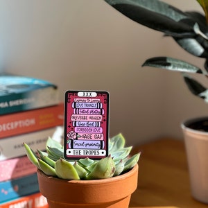 May include: A pink and black enamel pin with a stack of books that list common romance tropes, including "Enemies to Lovers", "Love Triangle", "Fated Mates", "Reverse Harem", "One Bed", "Forbidden Love", "Age Gap", and "Forced Proximity". The pin is sitting on top of a potted succulent.